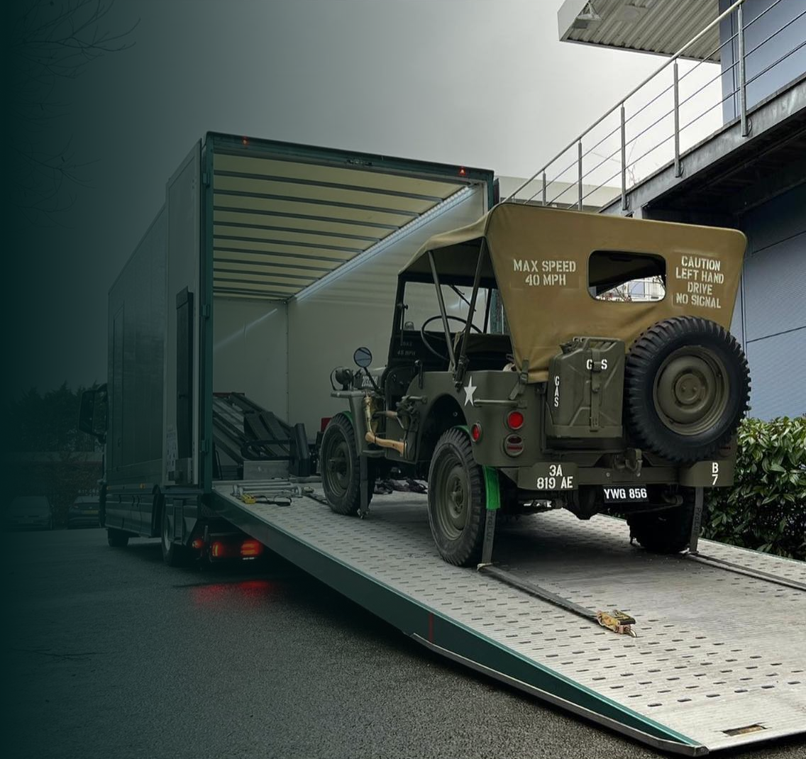 Vintage military jeep being loaded onto a large enclosed truck ramp outside a building.
