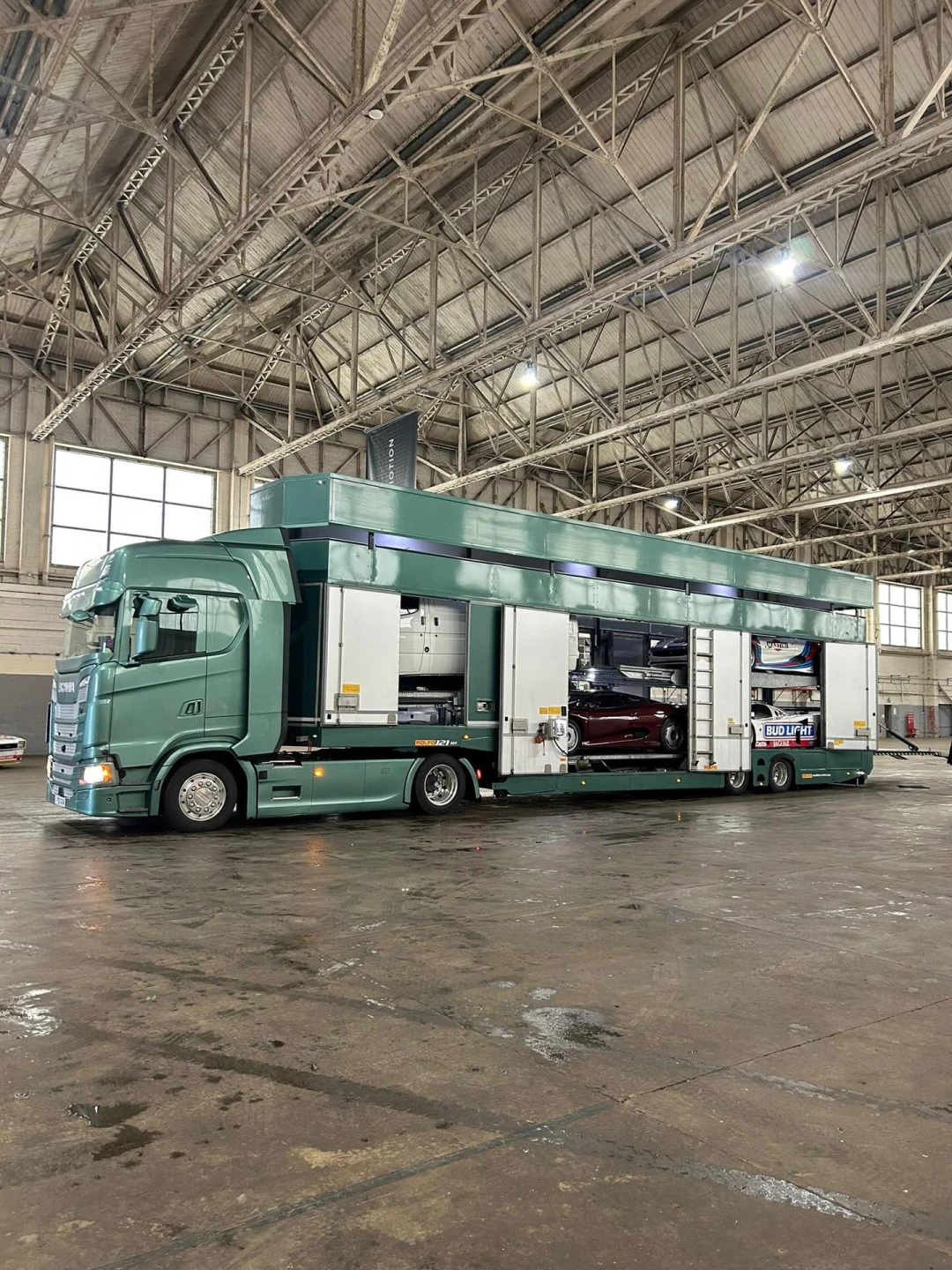 Vintage silver race car loaded on a green enclosed transport truck ramp in front of a building.