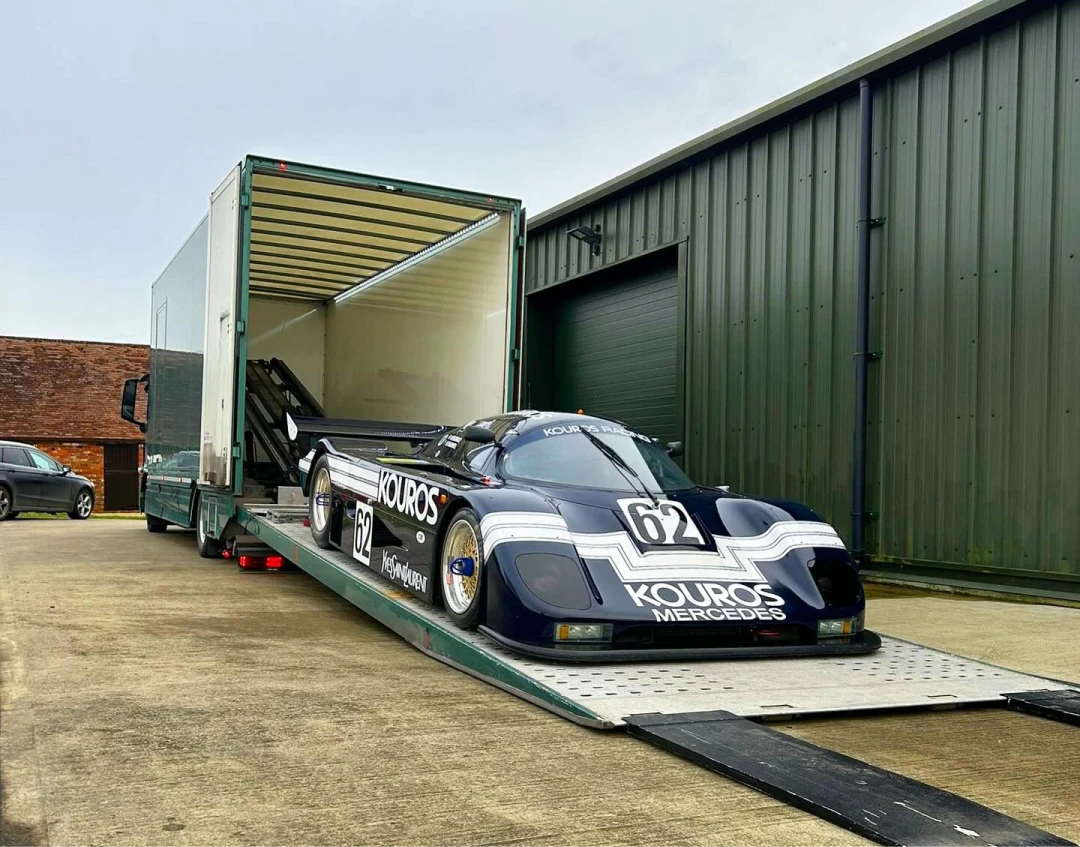 Vintage silver race car loaded on a green enclosed transport truck ramp in front of a building.
