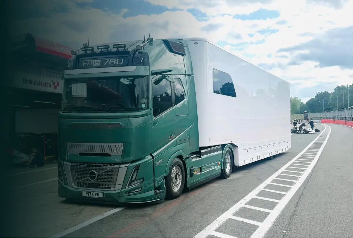 Green Volvo FH16 780 truck with white trailer parked on a racetrack pit lane near a pit stop building.