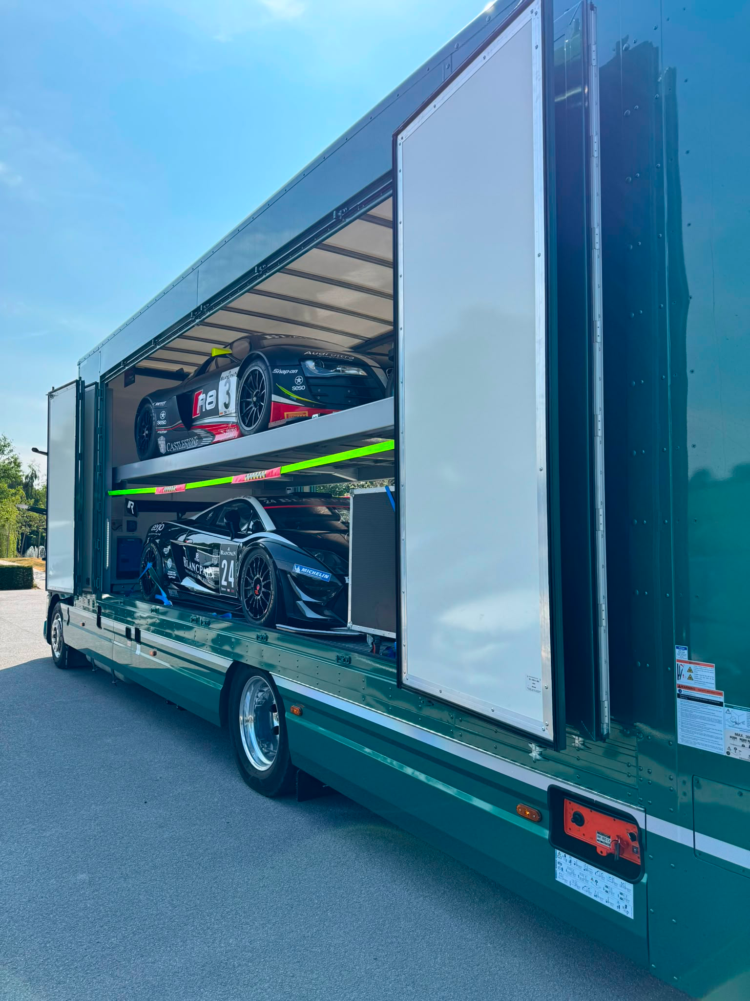Vintage silver race car loaded on a green enclosed transport truck ramp in front of a building.