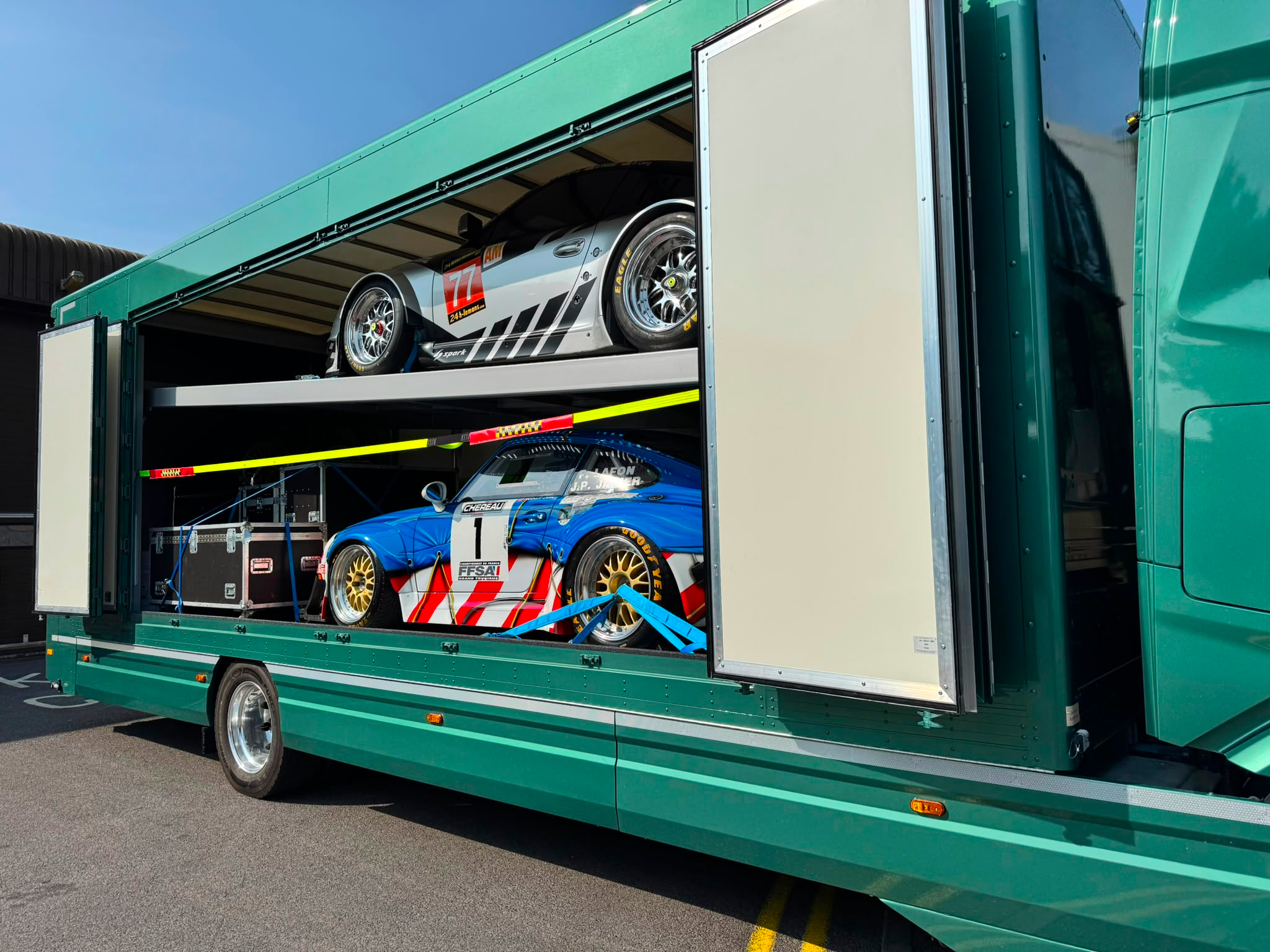 Yellow classic sports car being loaded into a large green enclosed car transport trailer on a concrete surface.