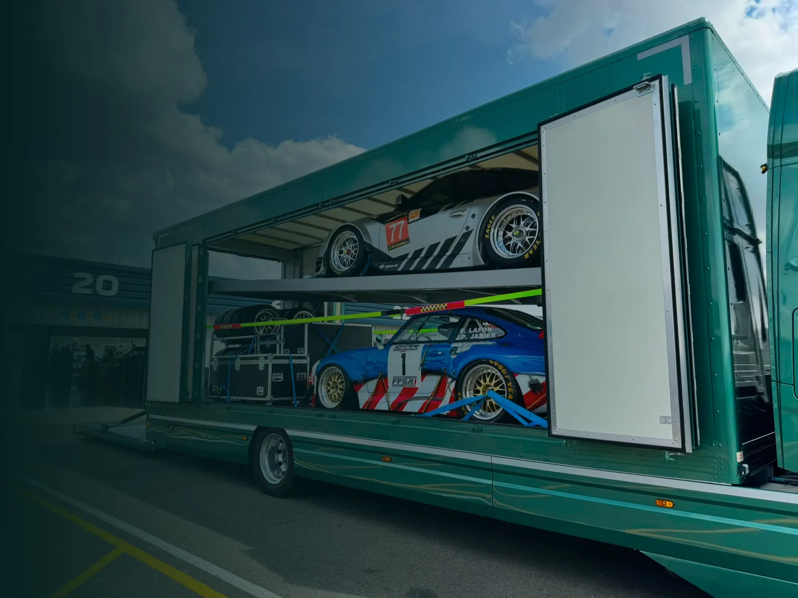 Two race cars loaded inside a green transport truck, one on an upper rack and one below it.