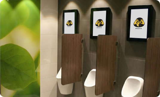 Men's restroom urinals with wooden dividers and digital screens above each displaying a yellow hands logo with the word MEDIA, green leaf mural on left wall.