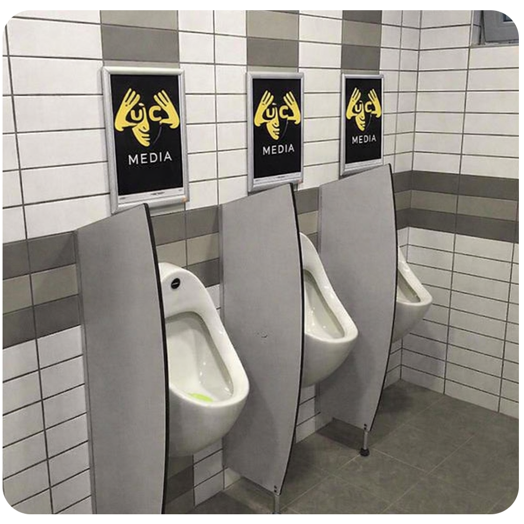 Three white urinals with gray privacy dividers mounted on a tiled wall, each under a framed poster showing hands making symbols and the word MEDIA.