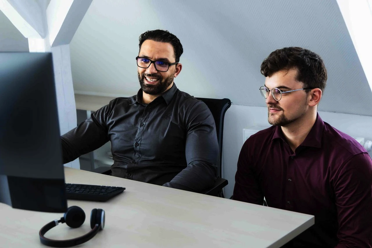 Zwei Männer mit Brillen sitzen an einem Schreibtisch und schauen gemeinsam auf einen Computerbildschirm.