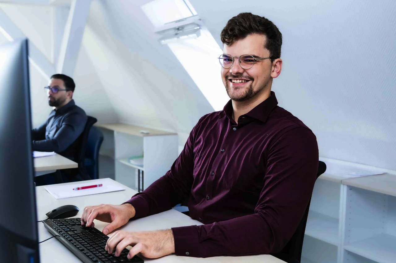 Lächelnder Mann mit Brille, der an einem Computer in einem modernen Büro arbeitet.