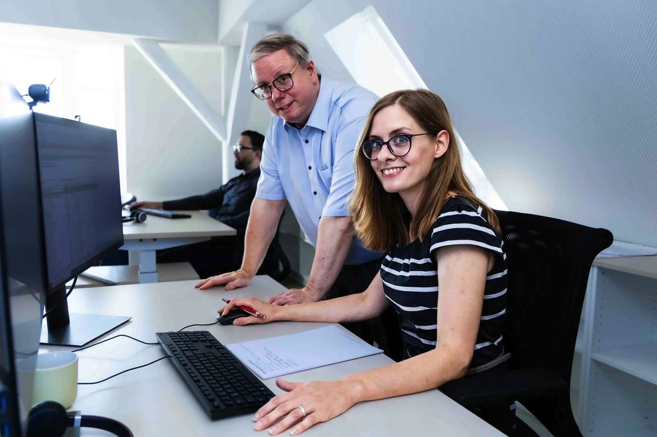 Frau mit Brille am Computerarbeitsplatz, ein älterer Mann steht neben ihr und beobachtet den Bildschirm.