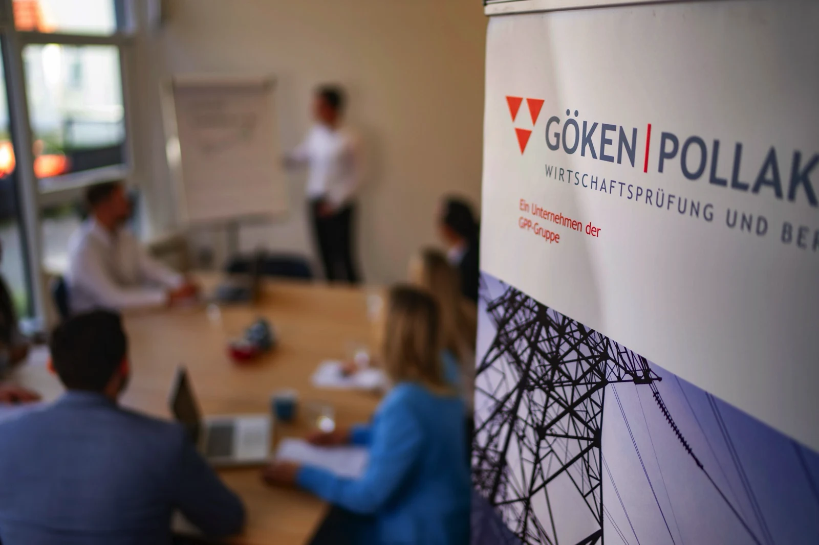 Besprechung in einem Konferenzraum mit fünf Personen an einem Tisch und einem Redner an einem Flipchart, daneben ein Banner der Wirtschaftsprüfungsgesellschaft Göken Pollak.