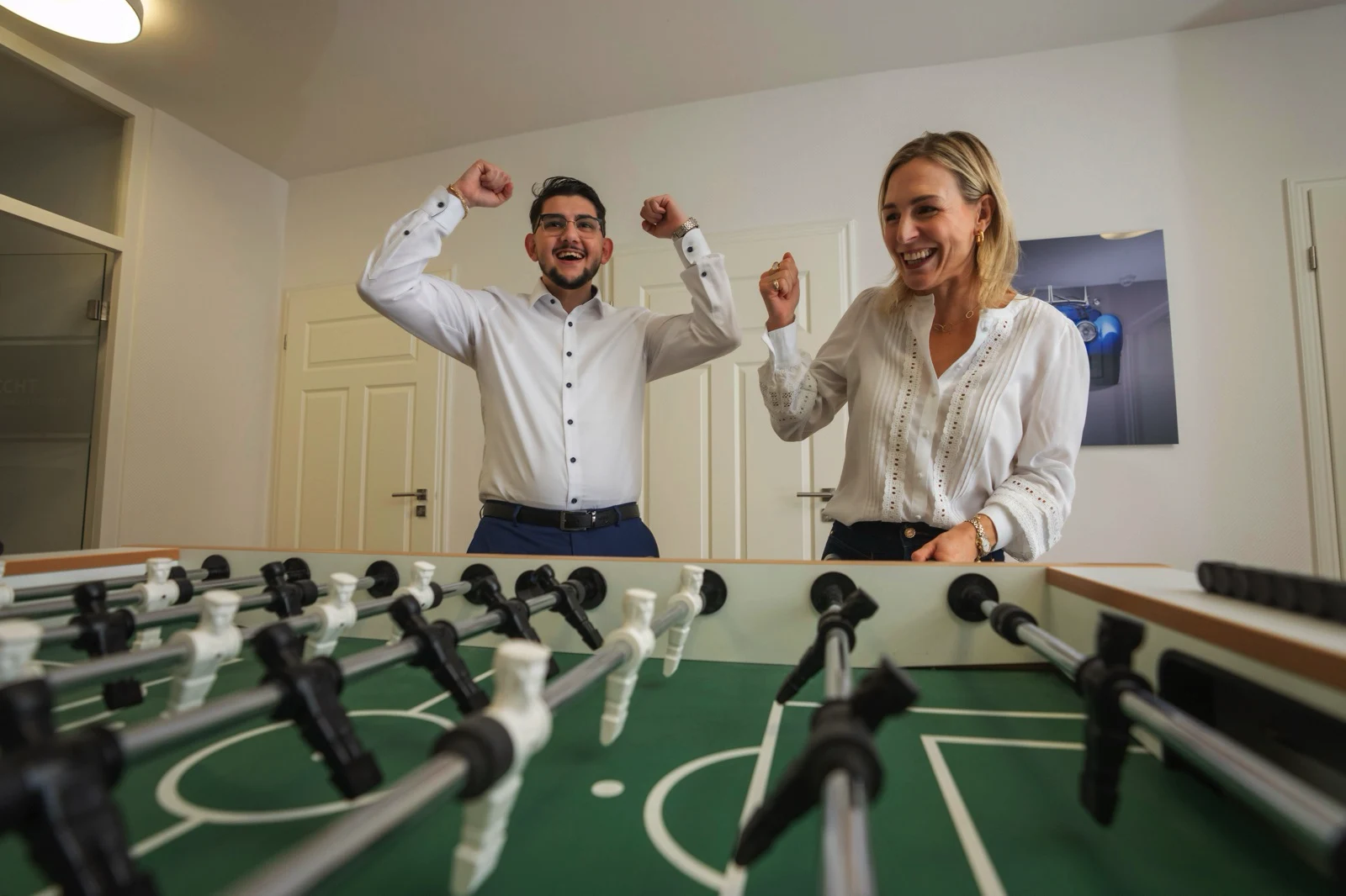 Zwei Erwachsene jubeln stehend hinter einem Tischfußballspiel in einem Raum.