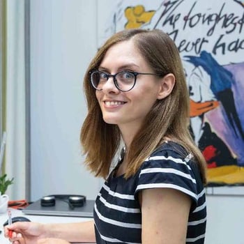 Junge Frau mit Brille und gestreiftem Shirt lächelt im inneren Raum vor einer farbenfrohen Wandkunst.
