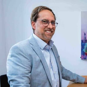 Mann mit Brille und grau-blauem Sakko lächelt in einem hellen Raum.
