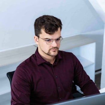 Junger Mann mit Brille und dunkelrotem Hemd, der konzentriert vor einem Computer sitzt.