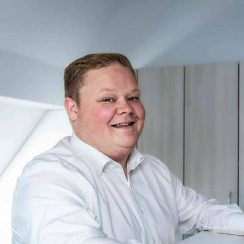 Lächelnder Mann in weißem Hemd vor einem Schrank in einem hellen Raum.