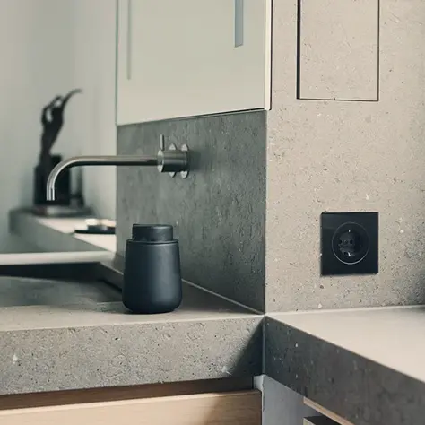 Moderne badkamer met een grijze stenen wastafel, wandkraan en een zwarte elektrische stopcontact.