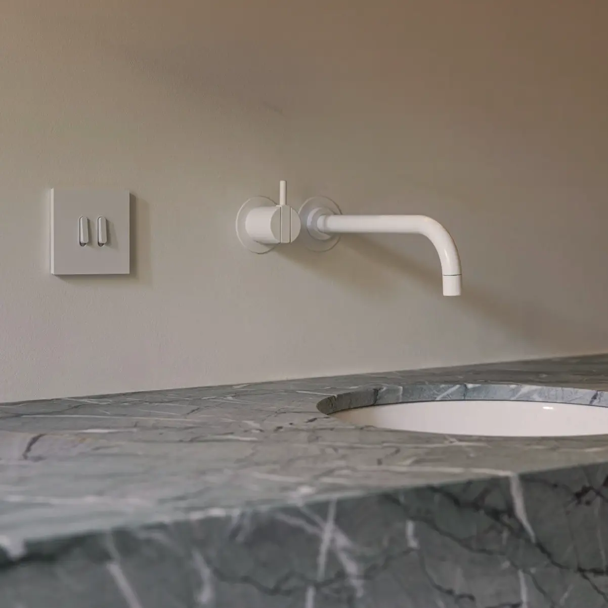 Moderne witte wandkraan boven een wastafel met een grijs marmeren blad en twee witte schakelaars op een beige muur.