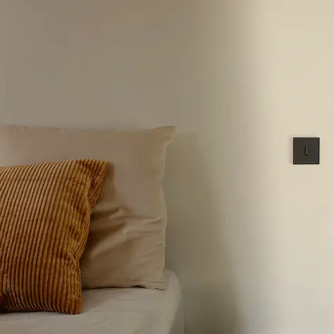 Een beige bed met een beige kussen en een bruin ribbelkussen tegen een lichte muur naast een zwarte lichtschakelaar.