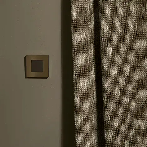 Beige gordijn naast een lichtschakelaar op een grijze muur.