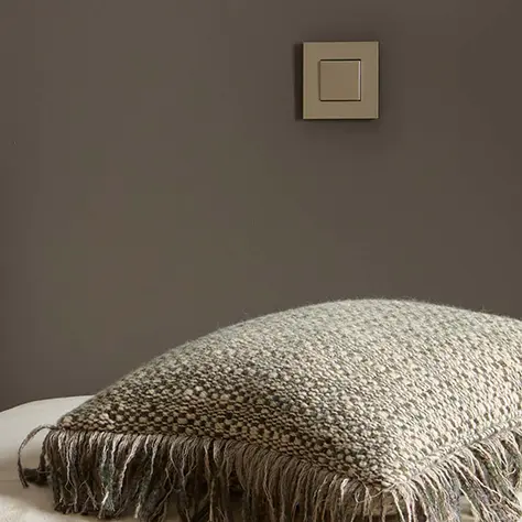 Beige gebreide kussen met franjes op een bruine achtergrond en een lichtschakelaar aan de muur.