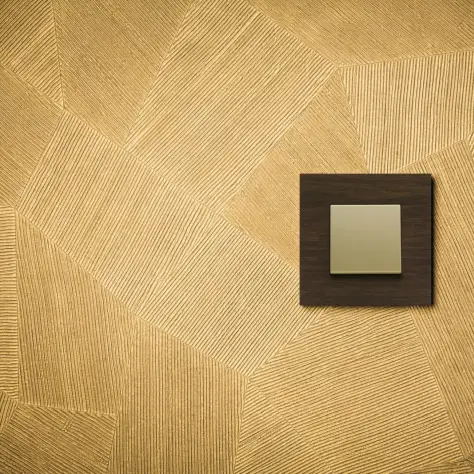 Vierkante lichtschakelaar op een goudkleurige muur met reliëfpatroon van lijnen in verschillende richtingen.