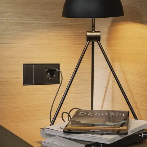 Zwarte metalen tafellamp met drie poten naast een zwart stopcontact op een houten muur, met een stapel boeken en een tijdschrift met de titel 'DESIGN' eronder.