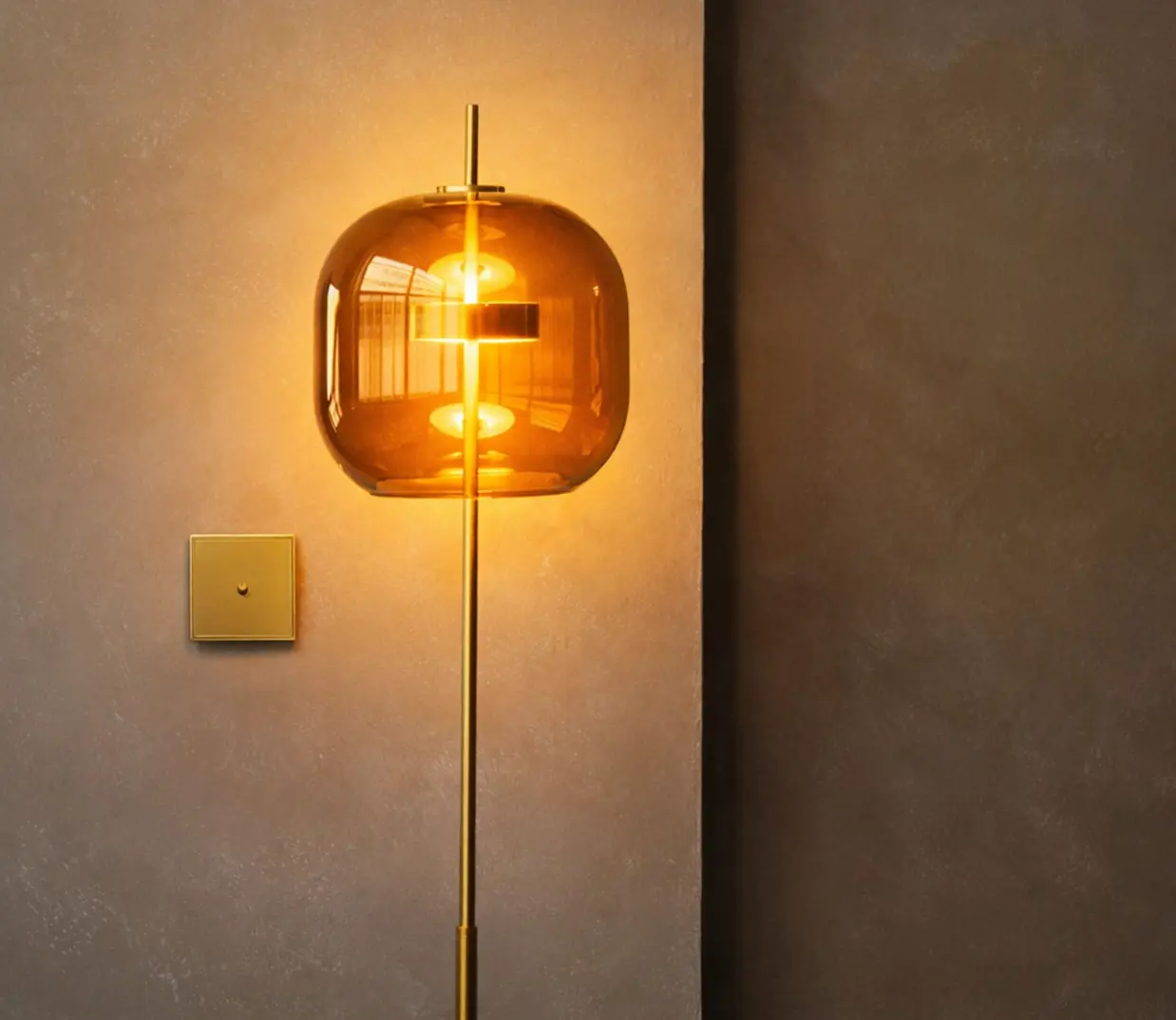 Moderne vloerlamp met een , bruine rookglas kap met een mat messing poot, voor een lichtbruine muur met een vierkante messing design schakelaar.