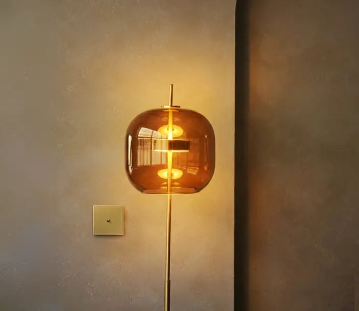 Moderne vloerlamp met een ronde, bruine glazen kap aan een matte gouden standaard voor een lichtbruine muur met een vierkante schakelaar.