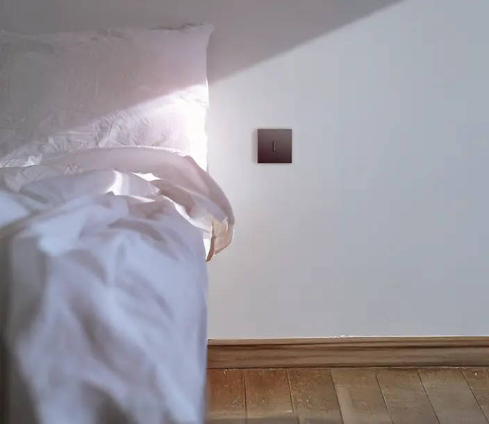 Witte beddengoed op een bed naast een witte muur met een zwarte lichtschakelaar en houten vloer.