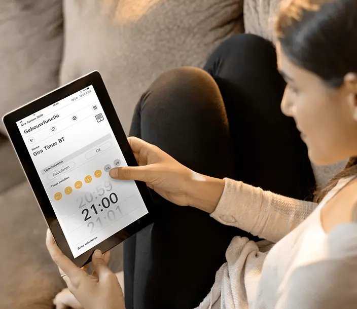 Vrouw zit relaxed op de bank en over haar schouder zien we dat ze smart-home bedient op haar tablet