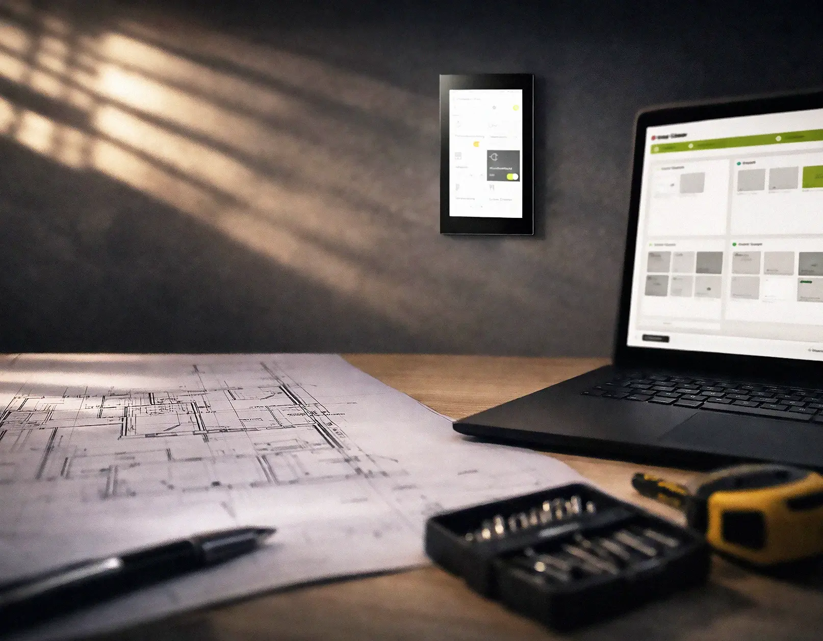Een duistere scene met zonlicht door de luxaflex en op tafel een bouwtekening, een laptop, een rolmaat en een pen met op de wand een Gira G1 smart-home bedieningspaneel