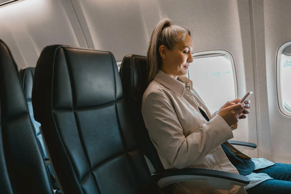 Woman sitting in an airplane seat by the window, looking at her phone.