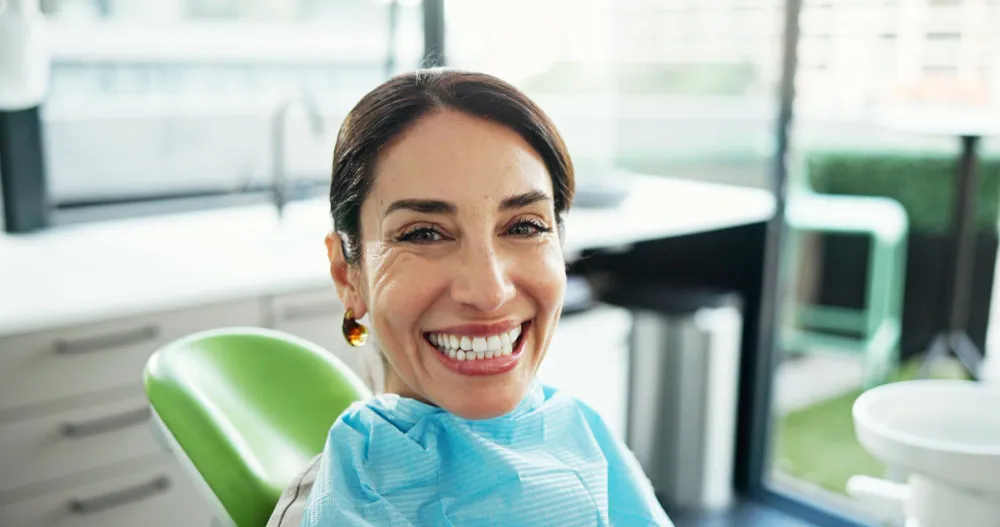 Smiling woman sitting in a green dental chair wearing a blue dental bib in a bright dental clinic.
