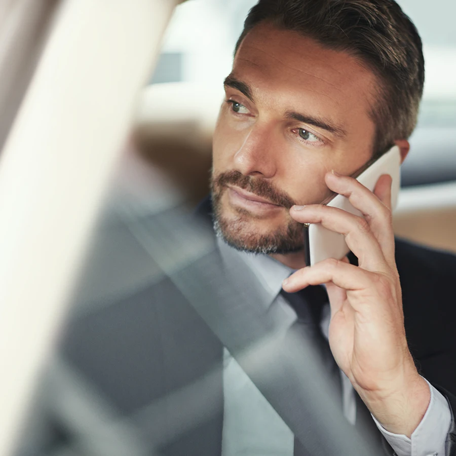 Man in business attire sitting in a car, talking on a smartphone and looking out the window.