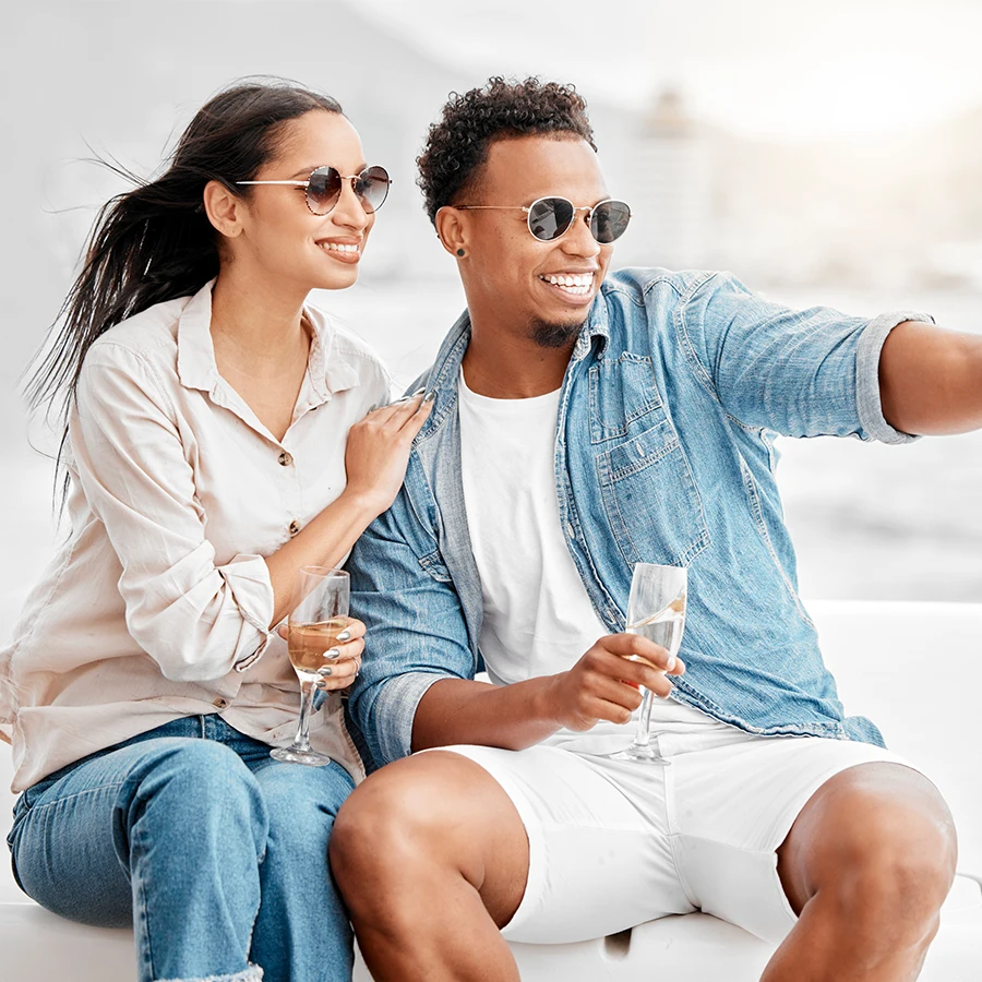 Smiling couple wearing sunglasses sitting close together, holding champagne glasses, and taking a selfie.