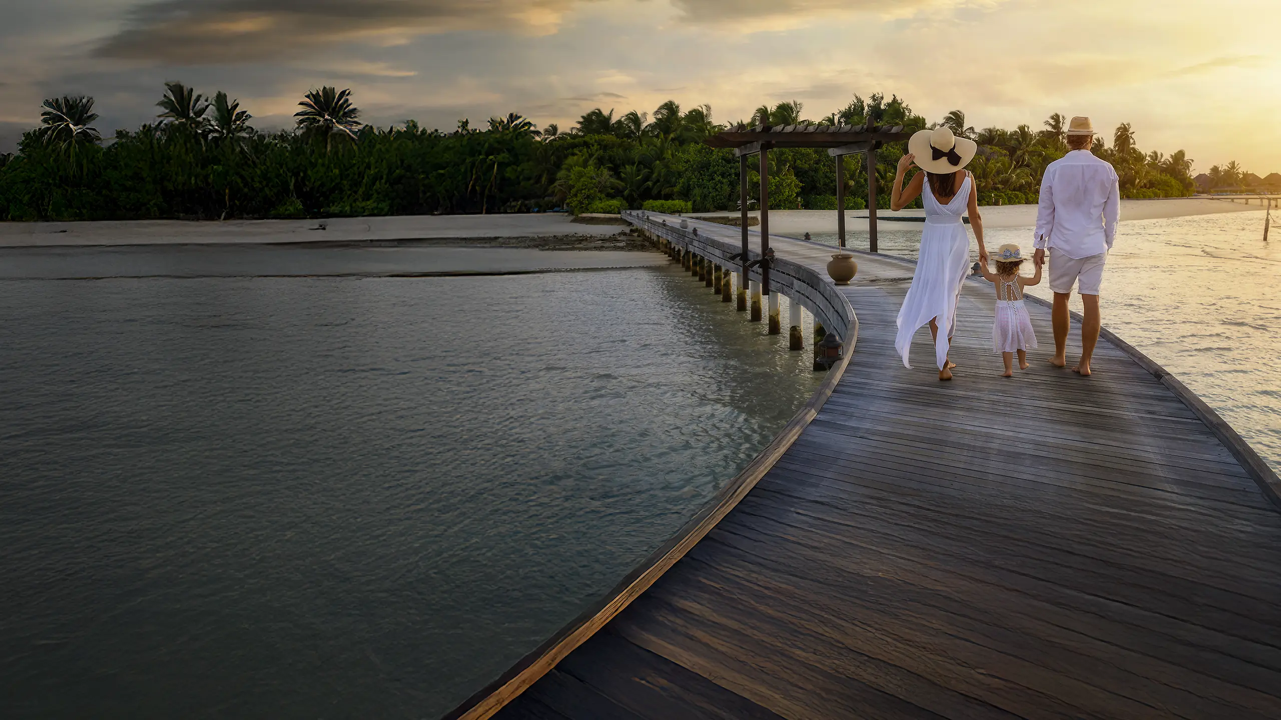 A family of three walking hand-in-hand on a wooden pier at sunset near tropical trees and calm water.