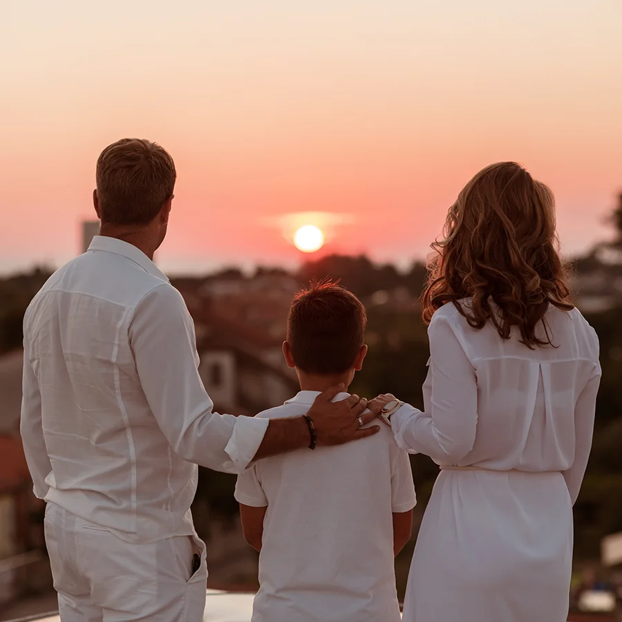 A family of three in white clothing watching the sunset together outdoors.