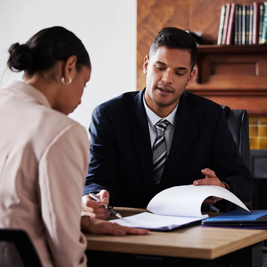Man in a suit explaining documents to a woman seated at a desk in an office setting.