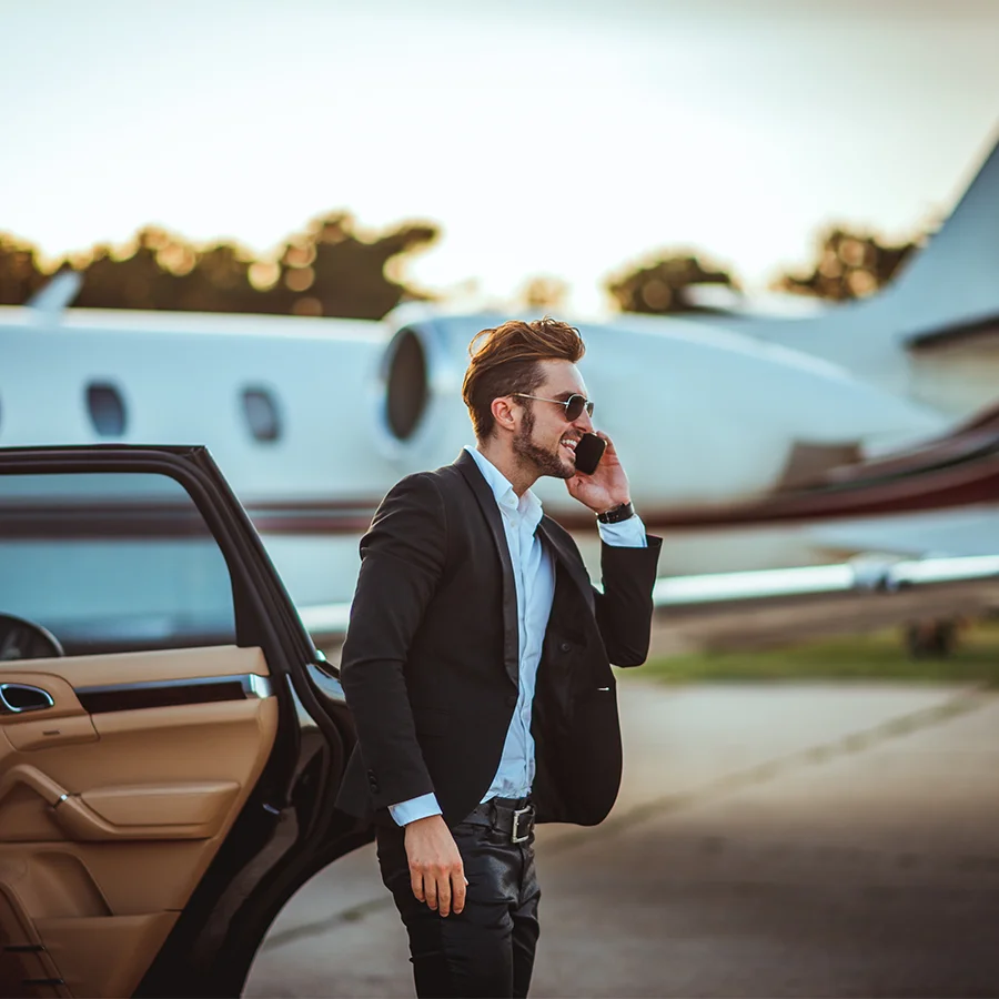 Man in sunglasses and suit talking on phone beside a car with an airplane in the background at sunset.