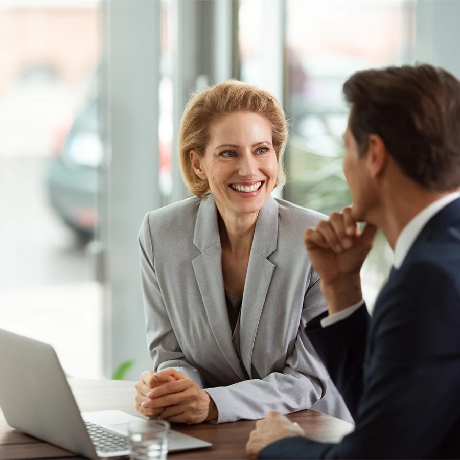 Smiling businesswoman in gray suit talking to a man in a navy suit at a desk with a laptop.