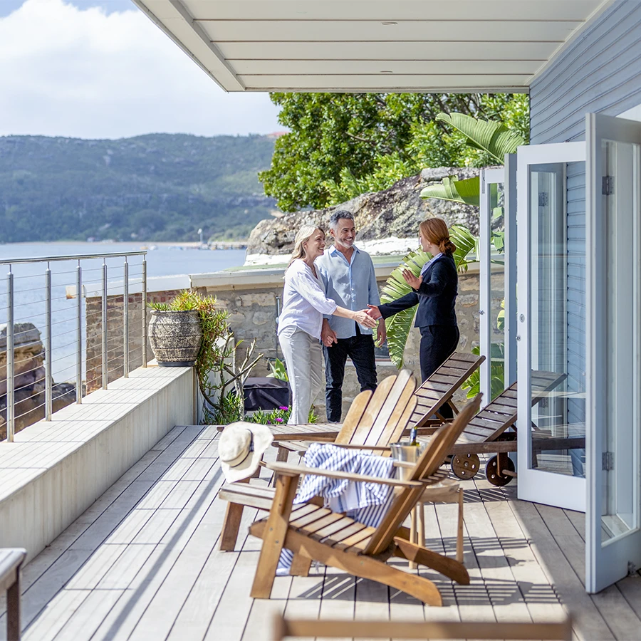 A couple shaking hands with a real estate agent on a wooden balcony overlooking water and hills.