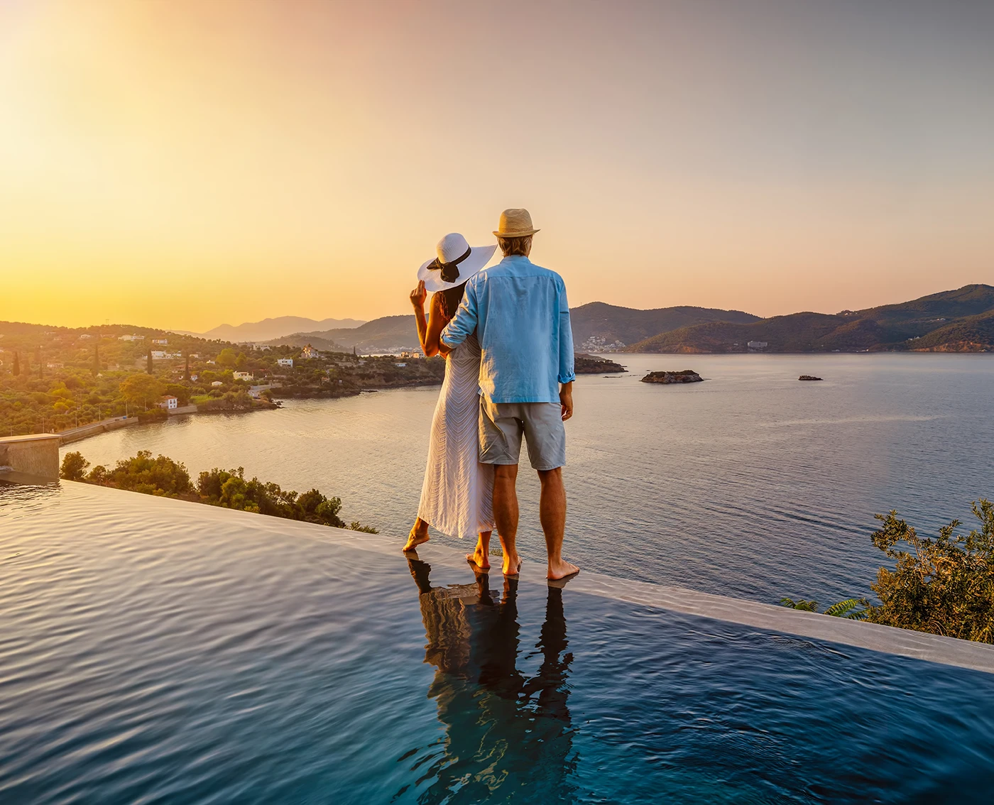 Couple standing barefoot at edge of infinity pool overlooking a calm sea and hills during sunset.
