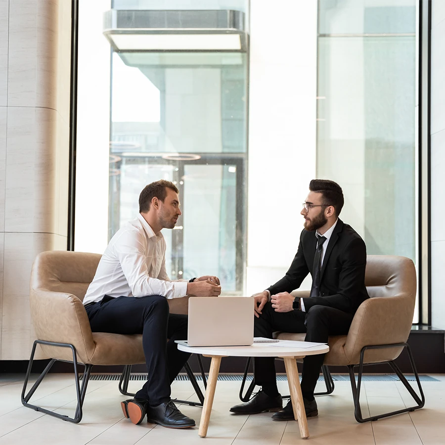 Two businessmen sitting in armchairs facing each other with a laptop on a small round table between them in a modern office.