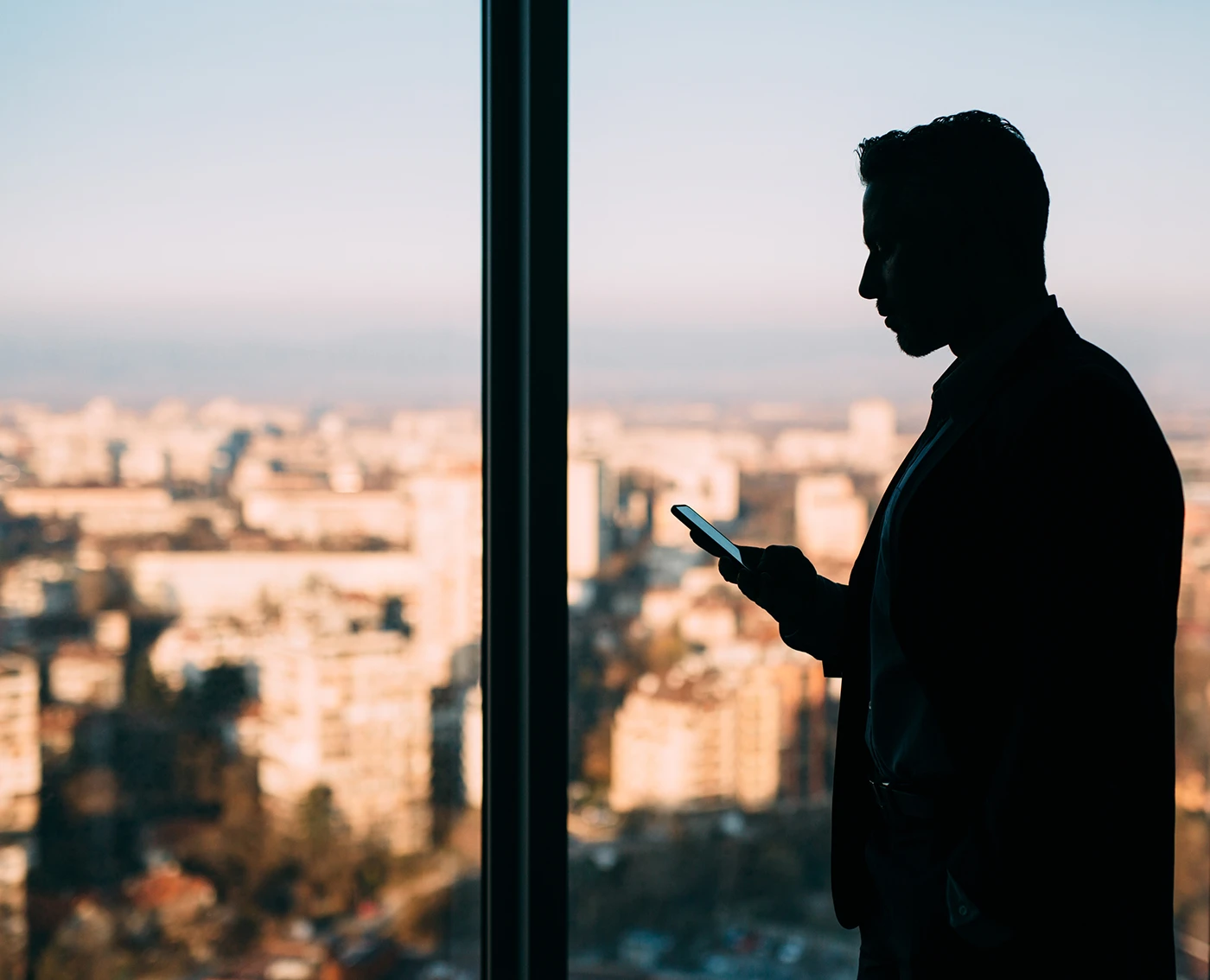 Silhouette of a man using a smartphone with a blurred cityscape visible through a large window in the background.