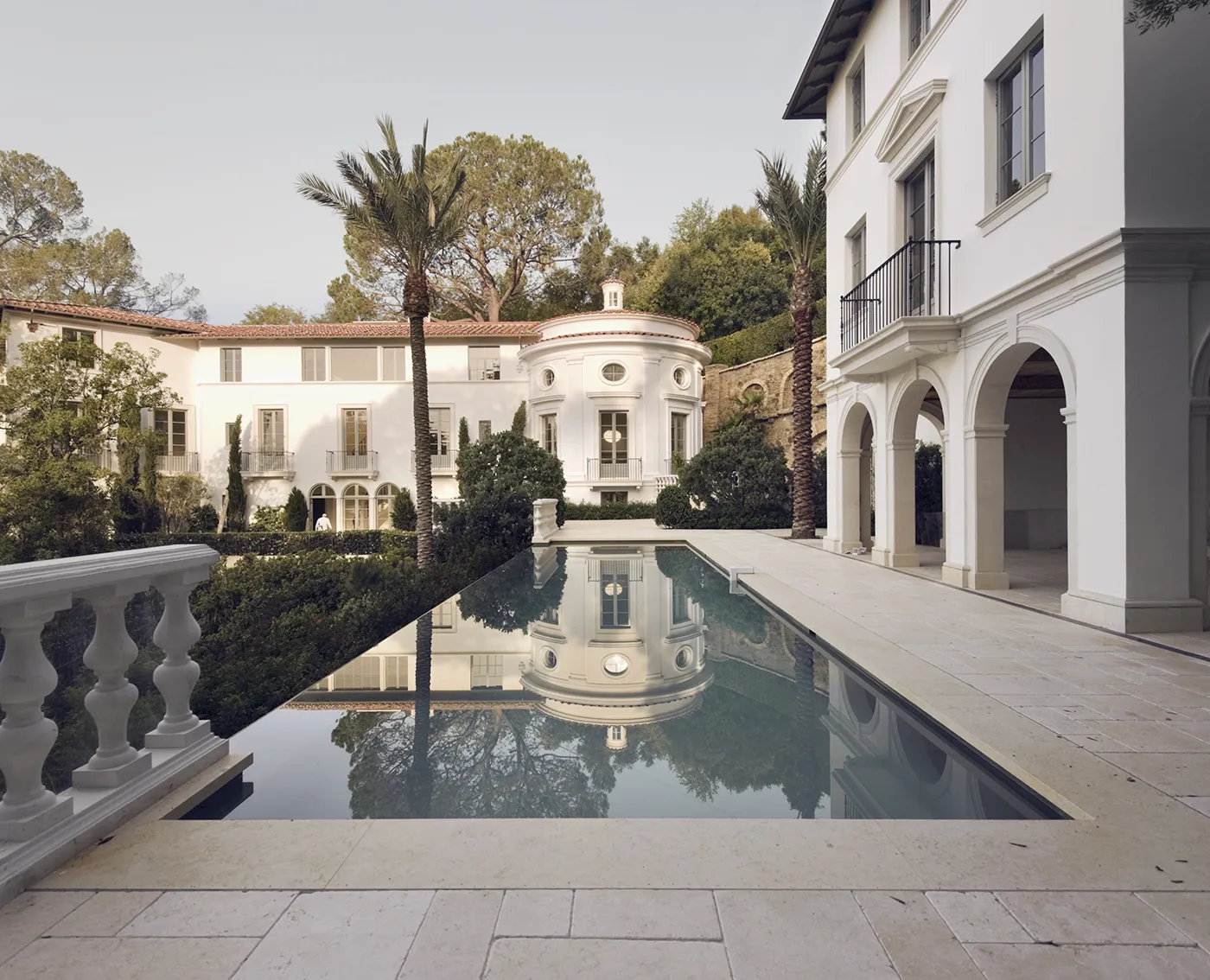 Elegant white mansion with arched balconies and a reflective rectangular pool surrounded by palm trees and greenery.