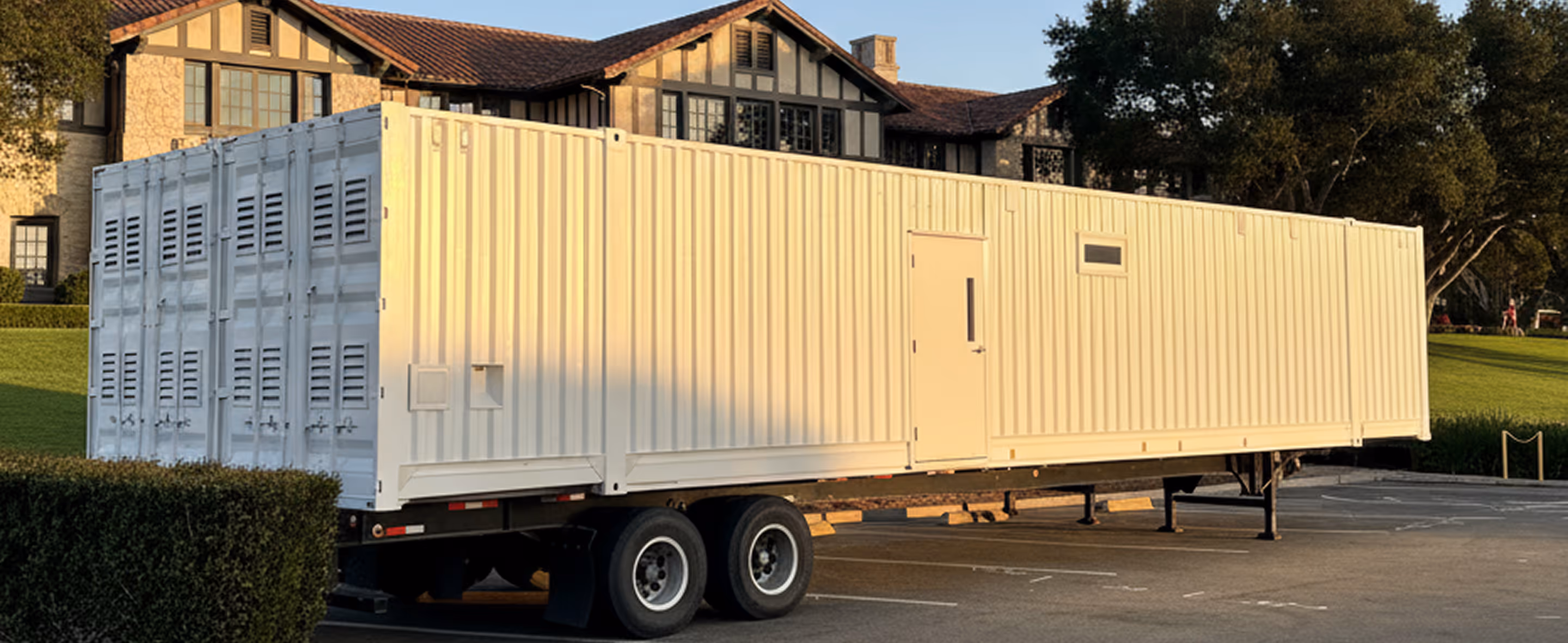 Large white mobile shipping container kitchen trailer parked near a green hedge in front of a Tudor-style building.