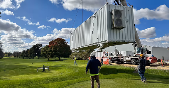 Construction workers guiding a large white shipping container kitchen being lifted by a crane on a grassy field with trees under a partly cloudy sky.