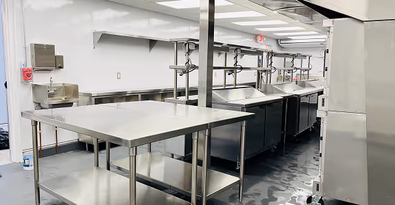 Clean commercial kitchen with stainless steel countertops, sinks, and work tables under fluorescent lights.