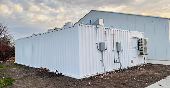 White shipping container kitchen with utility boxes and air conditioning unit placed outdoors on bare ground near a large industrial building under a cloudy sky.