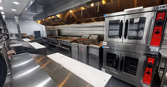 Clean commercial kitchen with stainless steel appliances including ovens, fryers, and grills under a ventilation hood.