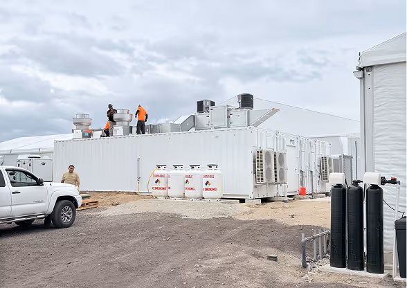 Three workers in orange safety gear on top of a large white industrial container with ventilation units and gas tanks labeled 'DANGER' and 'NO SMOKING' nearby, parked next to a white truck under a cloudy sky.